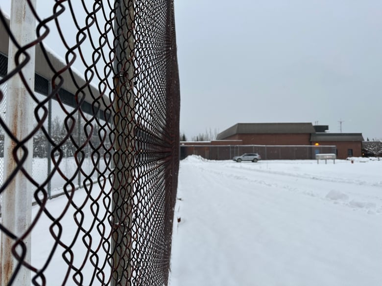 A large brick building is seen in the distance behind a chain link fence. There is snow on the ground.